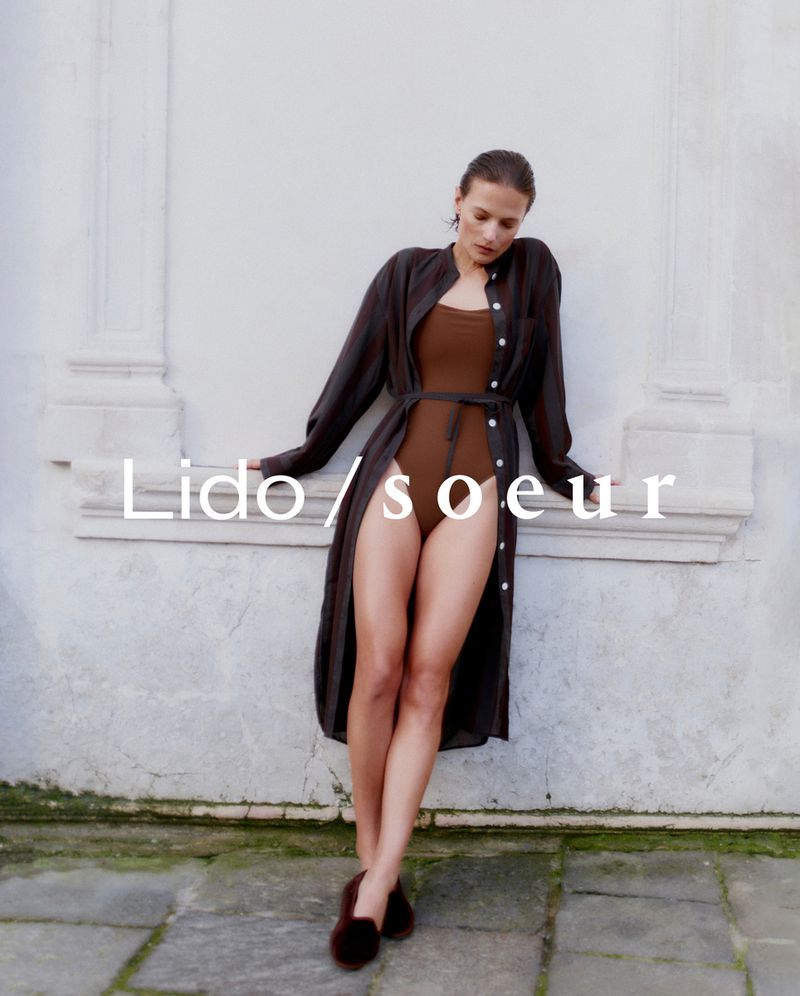 A woman in a brown bodysuit and a long dark shirt is leaning against a white wall, styled with dark flats, for a fashion shot.
