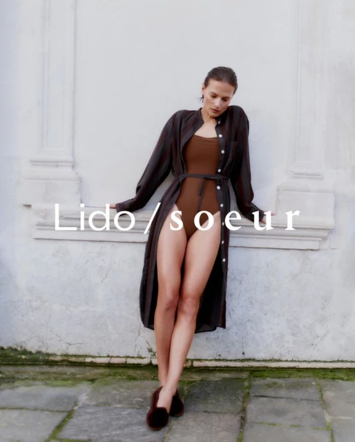 A woman in a brown bodysuit and a long dark shirt is leaning against a white wall, styled with dark flats, for a fashion shot.