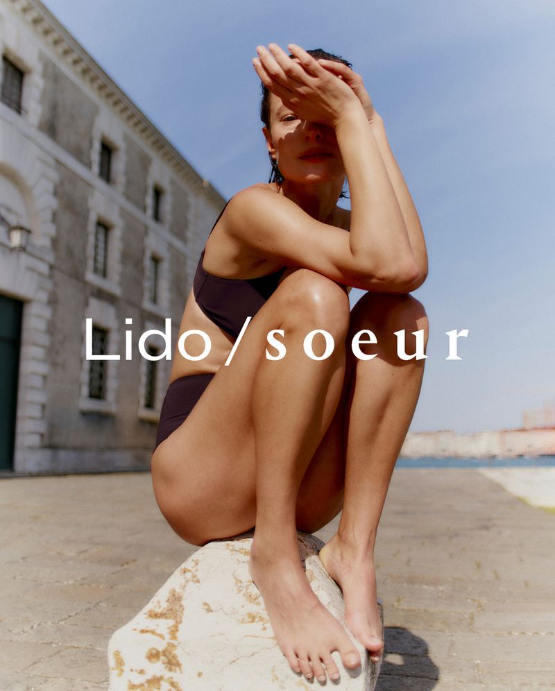 Woman in swimwear poses on a stone structure near a building and a body of water, with Lido/soeur text overlay. Fashion photography.