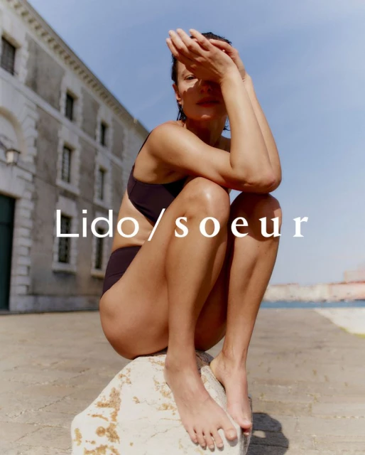 Woman in swimwear poses on a stone structure near a building and a body of water, with Lido/soeur text overlay. Fashion photography.