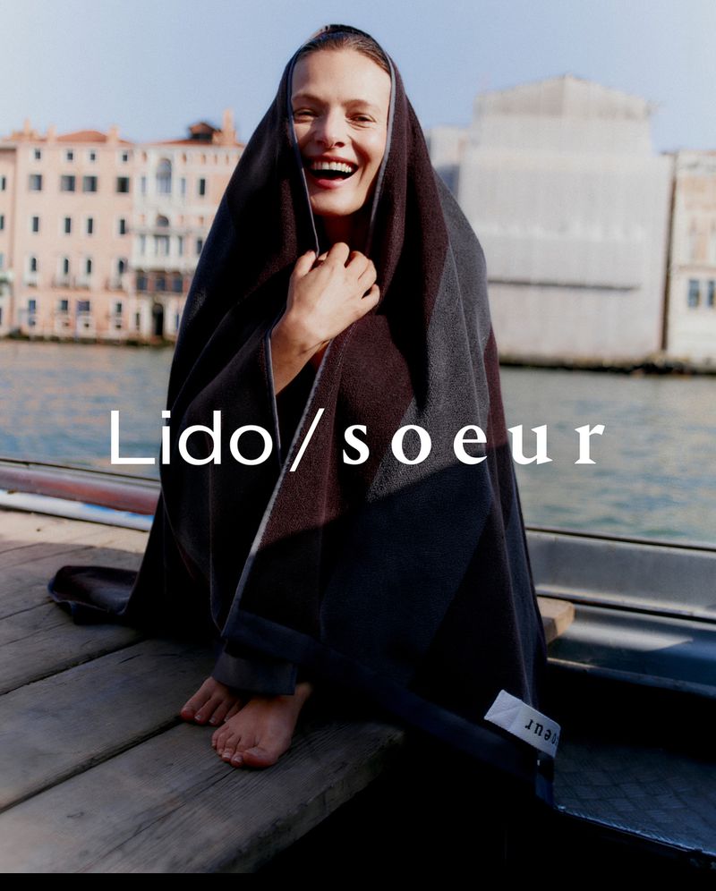 Adult woman wrapped in a blanket smiles on a dock in Venice, Italy. Buildings and water visible in background during daytime.