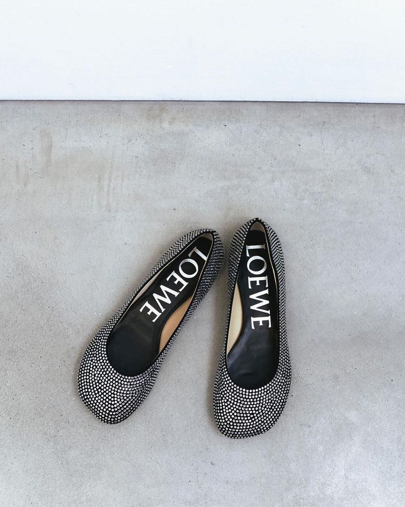 Pair of Loewe brand ballet flats with light-grey stone embellishments on a grey concrete surface, positioned below a white wall.