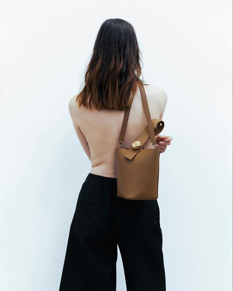 Back view of a slender topless woman wearing black culottes and a tan bucket bag with a crossbody strap against a white backdrop.