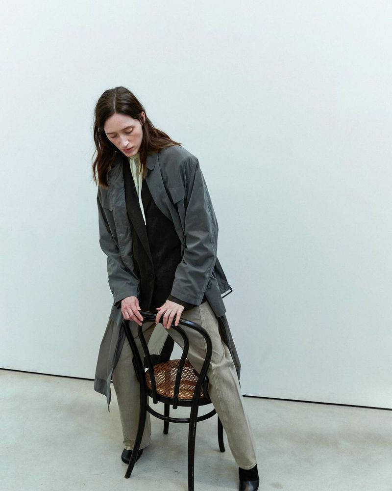 A slender woman in a gray trench coat sits on a black chair in a minimalist studio, posing for a fashion or editorial shot.