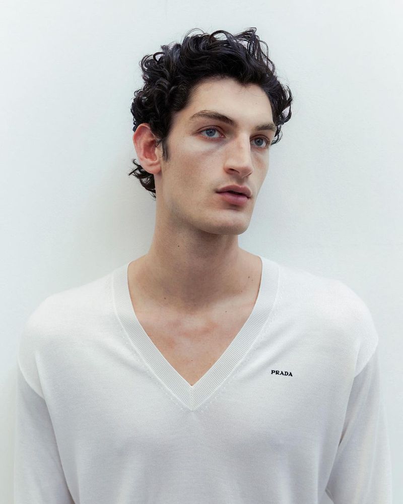 Close-up studio portrait of a young adult male with dark curly hair and light skin, wearing a white sweater with a V-neck.