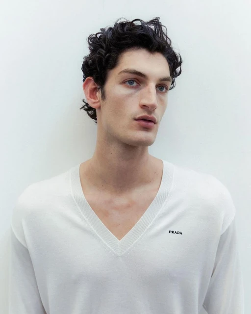 Close-up studio portrait of a young adult male with dark curly hair and light skin, wearing a white sweater with a V-neck.