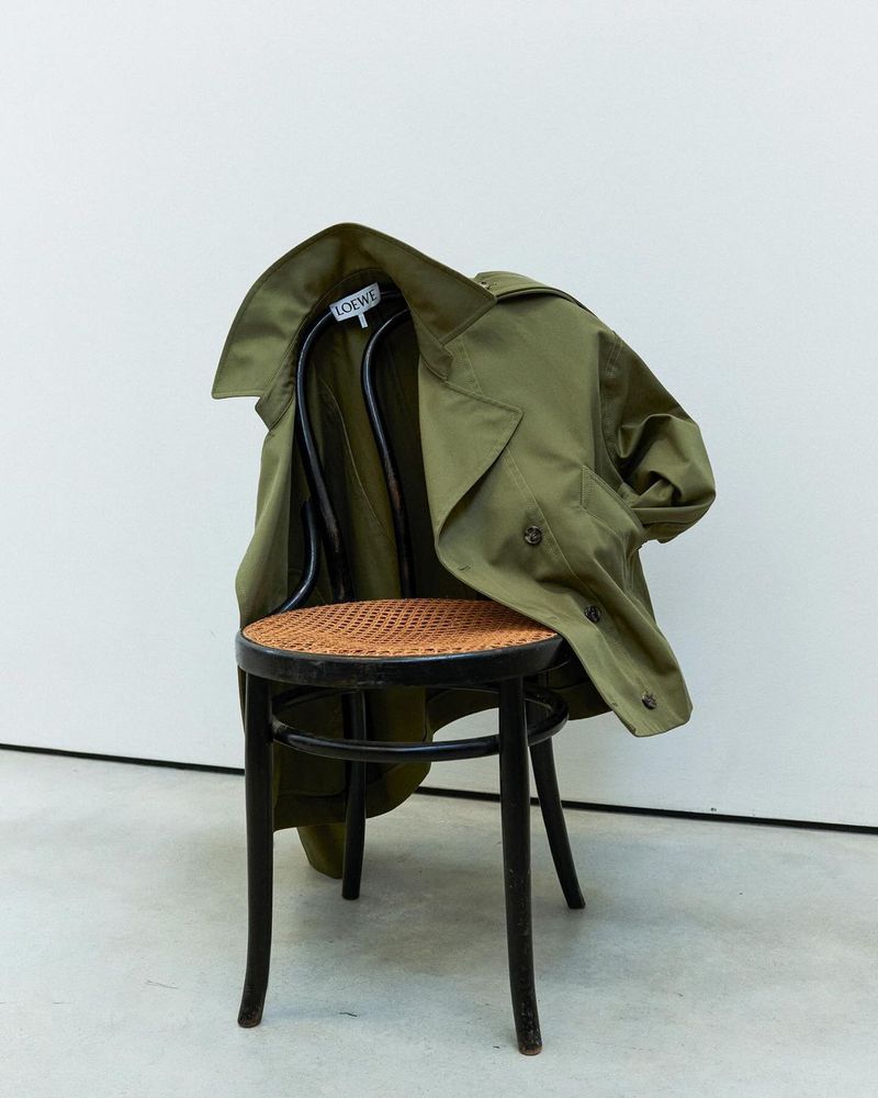 Olive green trench coat draped over a black wooden chair with a woven seat against a white wall in a softly lit studio.