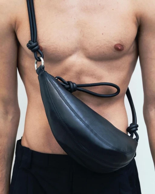 Torso shot of a muscular adult male wearing black trousers and a black leather shoulder bag with a round, modern design.