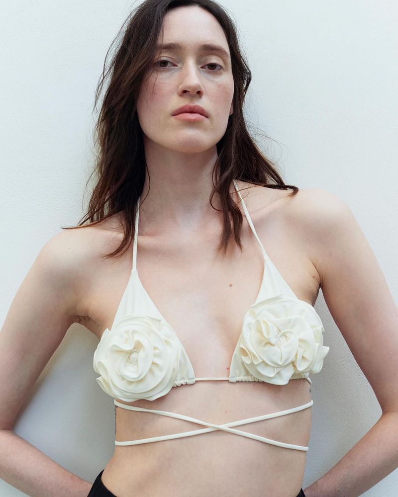 A woman poses in an off-white bikini top with floral details. The indoor portrait showcases a minimalist fashion aesthetic and natural light.