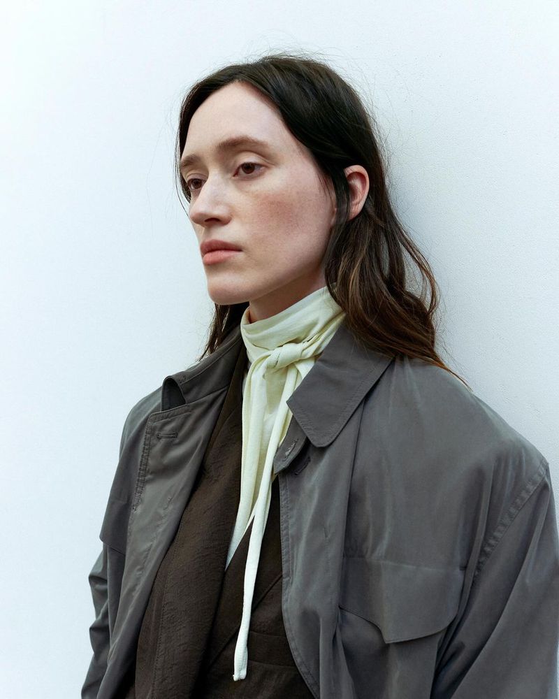A woman with long dark hair wears a grey trench coat and light-yellow scarf against a white background in a medium studio shot.