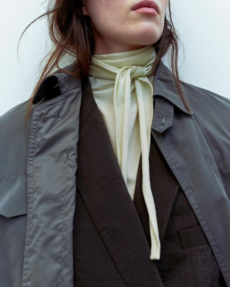 Fashion shot of a woman wearing a grey trench coat, black blazer, and cream-colored top tied at the neck, in front of a white background.