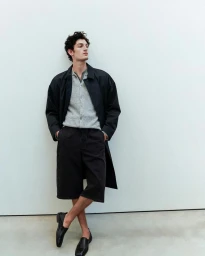 Model wearing dark coat, grey cardigan, and dark shorts against a plain white studio background
