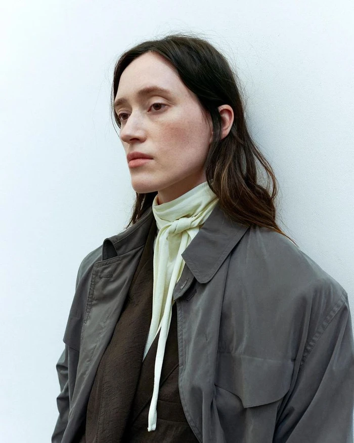 Model wearing a grey jacket and light scarf against a plain white wall