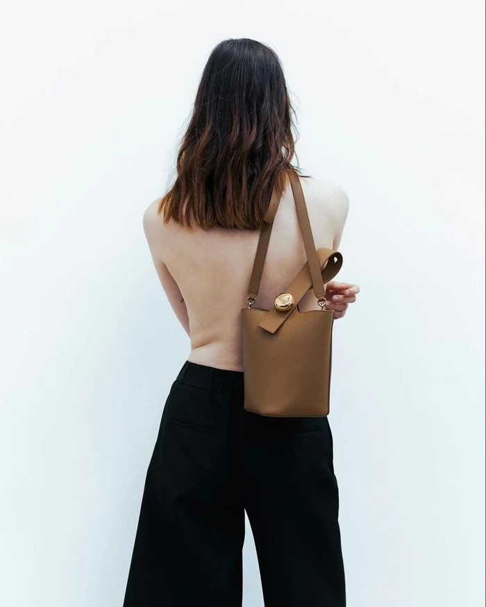 Model seen from behind wearing black trousers and holding a brown bucket bag against a white background