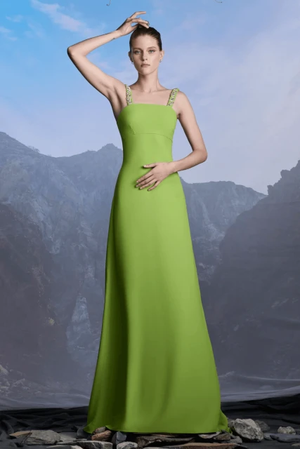 Woman poses in a long, light green dress with decorated straps against a grey mountain backdrop. The setting is stone.