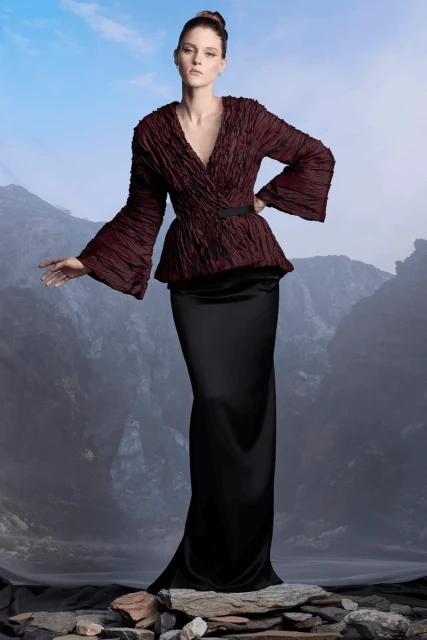 A woman in an elegant maroon top and black dress poses in a rocky outdoor setting, creating a fashion-forward and visually striking image.