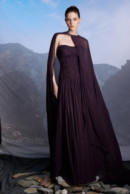 Adult female poses in a dark plum gown with cape over a mountain backdrop in an eye-level studio shot.