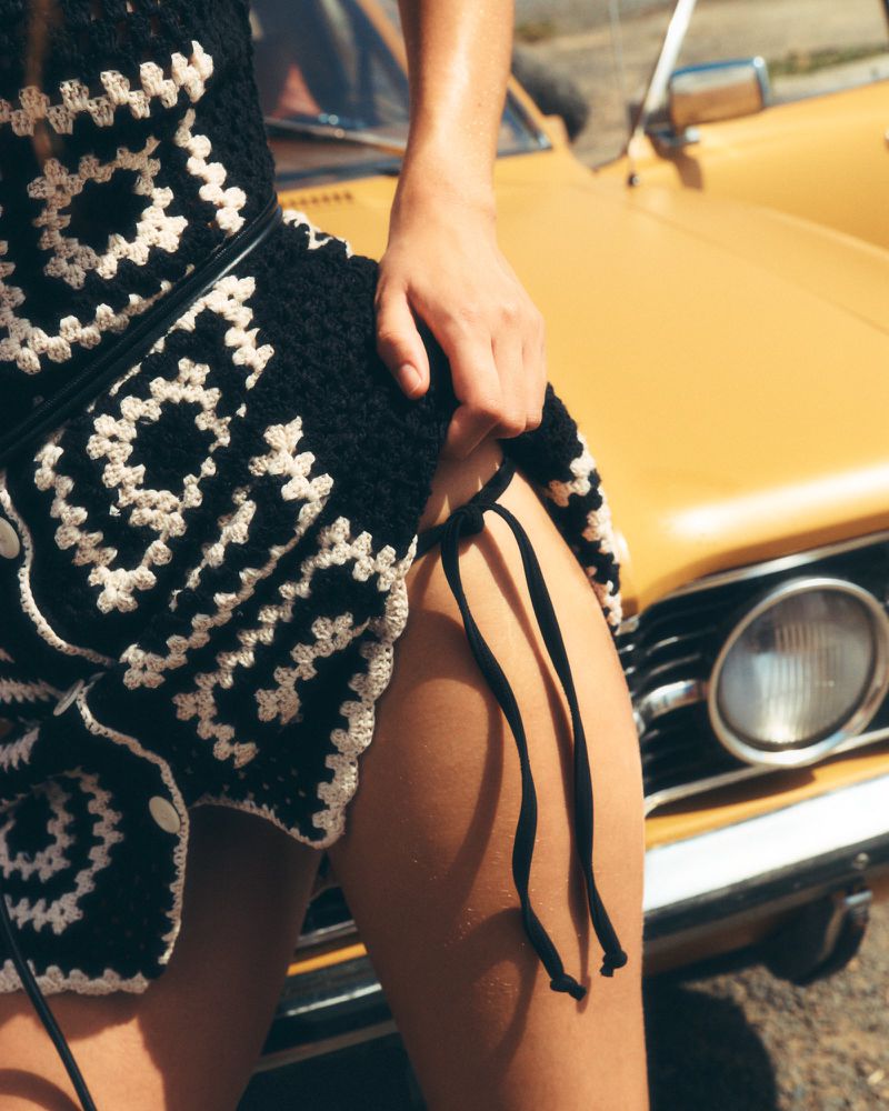 Close-up of an adult posing in a black and white crocheted dress with a yellow vintage car in the background on a sunny day.