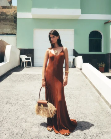 Woman in an orange maxi dress and holding a brown purse with fringe standing outdoors in front of a green building on a sunny day.