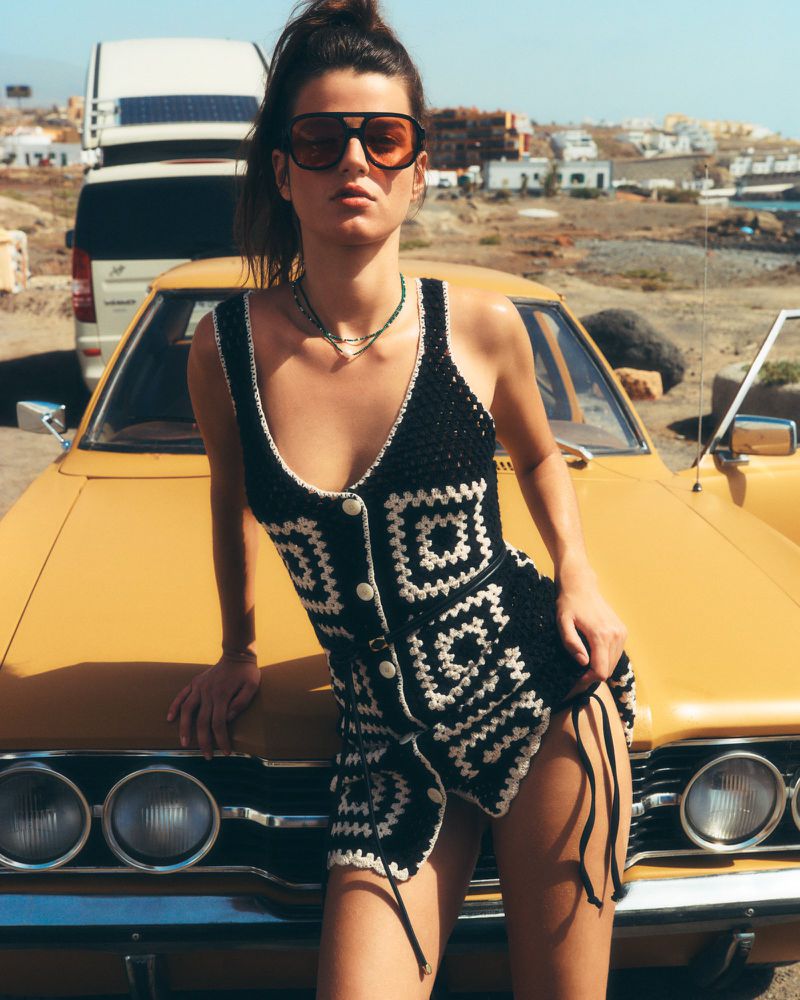 A woman in sunglasses poses against a classic yellow car on a sunny day. The scene includes a white van and buildings.