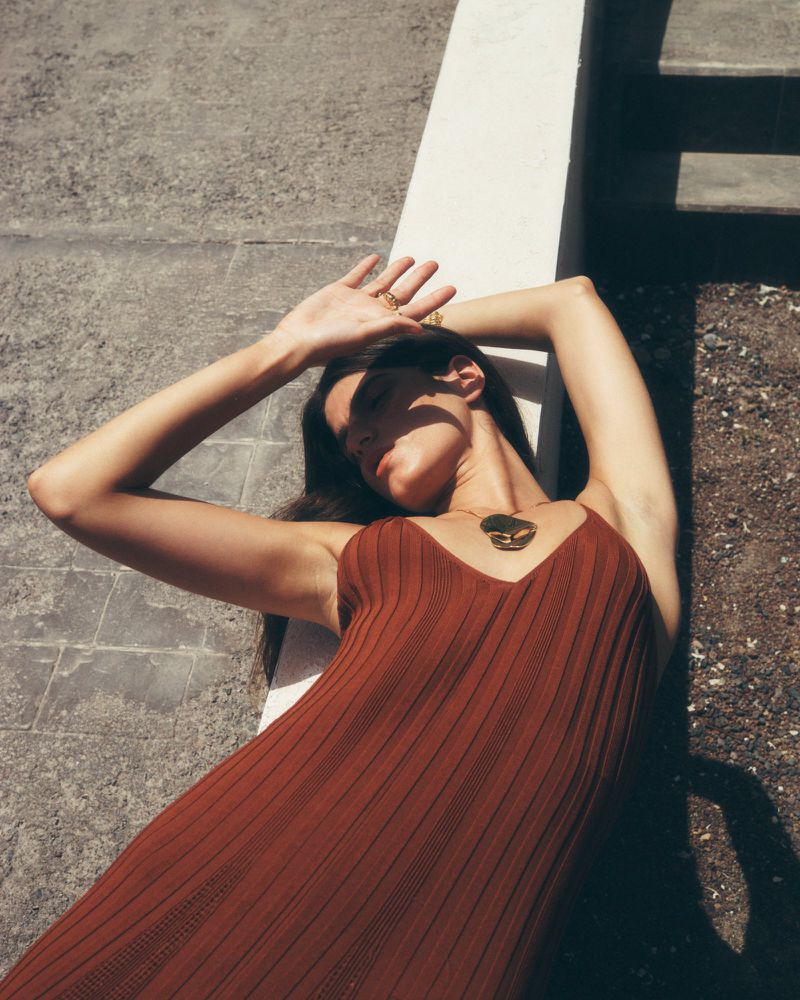 Woman in a brown dress lies in sunlight with her hand shading her face. She wears a necklace and ring. Outdoor setting. 