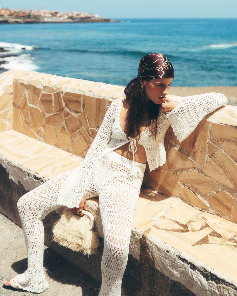 A woman in a crochet outfit sits on a stone wall with a purse, set against a backdrop of a blue sea and a sunny sky.