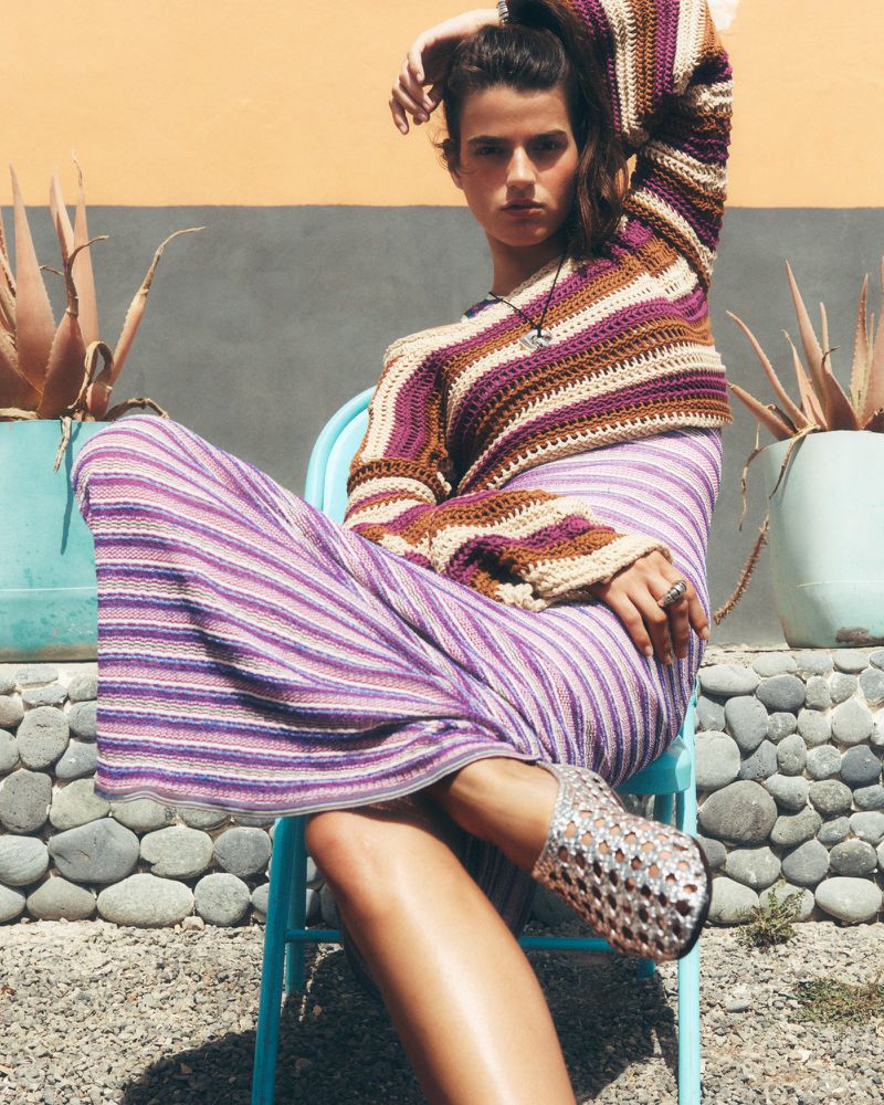 A woman in a striped top and skirt sits on a blue chair outdoors, styled fashionably, with a neutral wall and aloe plant in the backdrop.