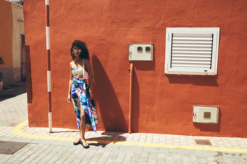 Young woman in a patterned crop top and wrap skirt poses against a vibrant orange wall in bright sunlight wearing sunglasses.