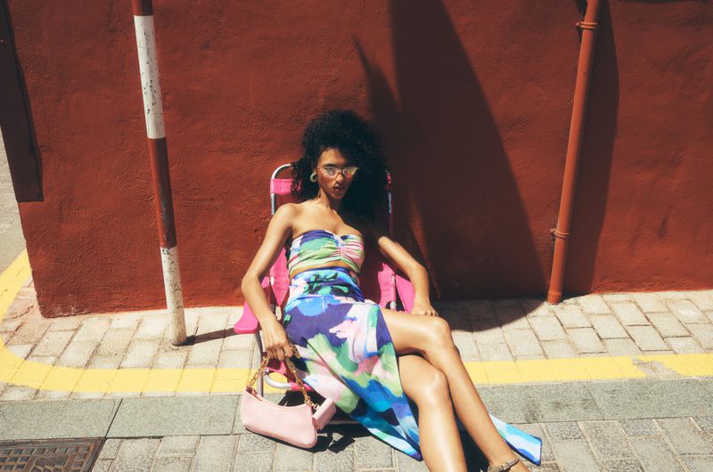 A woman poses in a floral skirt and top, seated on a pink folding chair outside on a sunny day with a reddish-brown background.