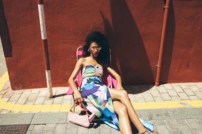 A woman poses in a floral skirt and top, seated on a pink folding chair outside on a sunny day with a reddish-brown background.