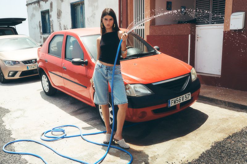 A woman in denim shorts washing an orange car with a hose outside near a building. There are other cars visible in background.