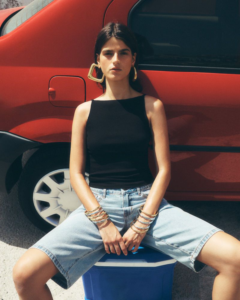 A brunette female fashion model sits on a blue cooler in front of a red car wearing a black top, light blue jean shorts, and gold earrings.