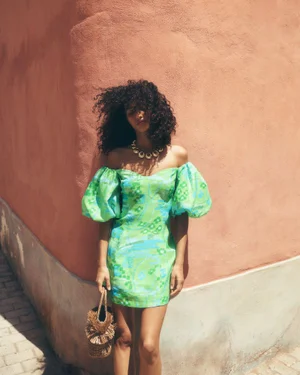Model in a green off-the-shoulder dress leaning against a textured peach wall, holding a woven bag