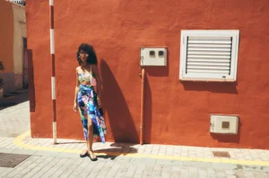 Model standing against a vibrant orange wall wearing a blue patterned two-piece outfit and sandals.
