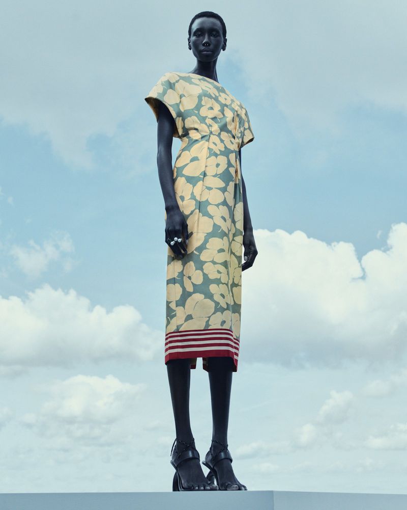 A full shot of an adult fashion model posing in a floral dress and black high heel shoes against a backdrop of blue sky and clouds.