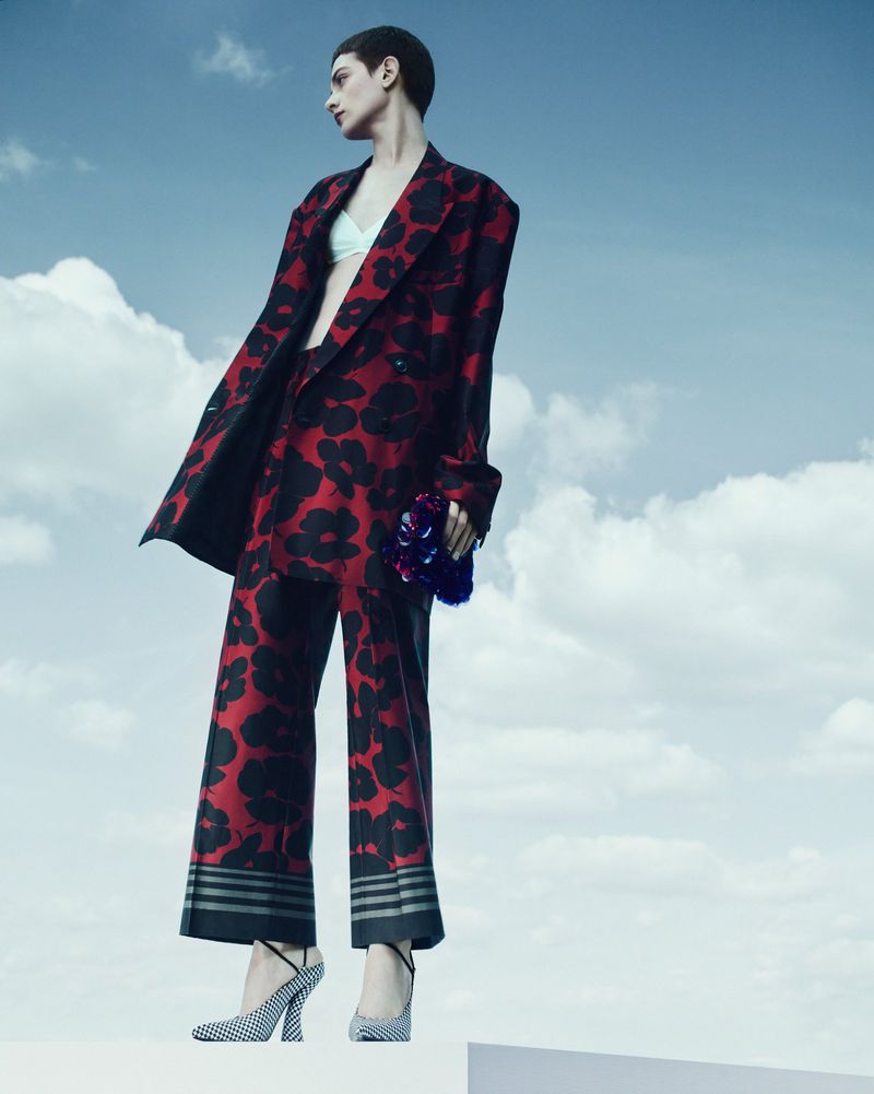Woman in floral print suit posing outdoors against a cloudy sky. She is wearing patterned high heels and holding a small bouquet of flowers.
