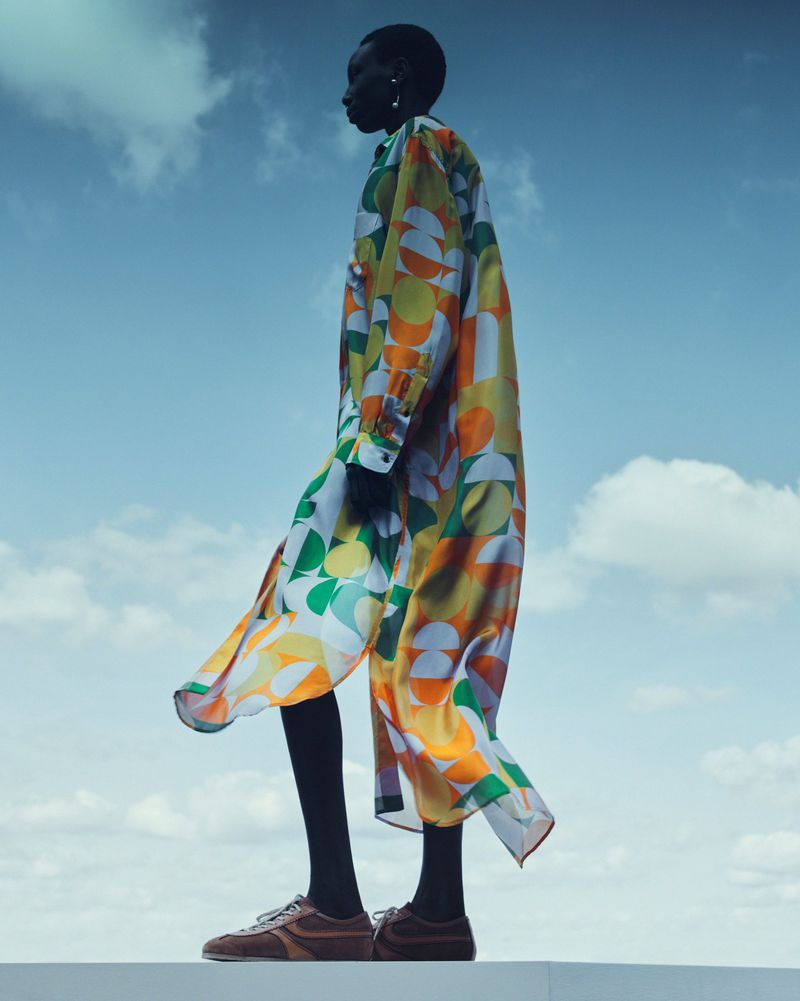 A slender Black woman stands in profile wearing an oversized patterned shirt dress, brown sneakers, and black leggings against a sky background.