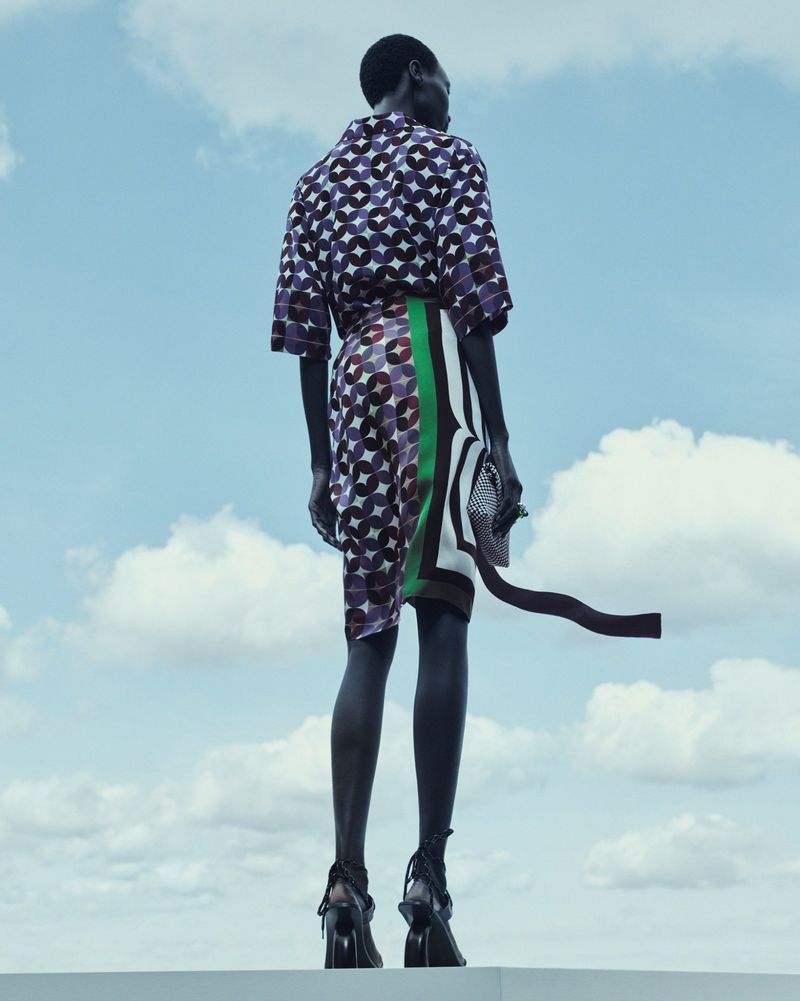 Fashion photo of an adult woman in a patterned top and skirt standing against a blue sky with clouds, shot from a low angle.