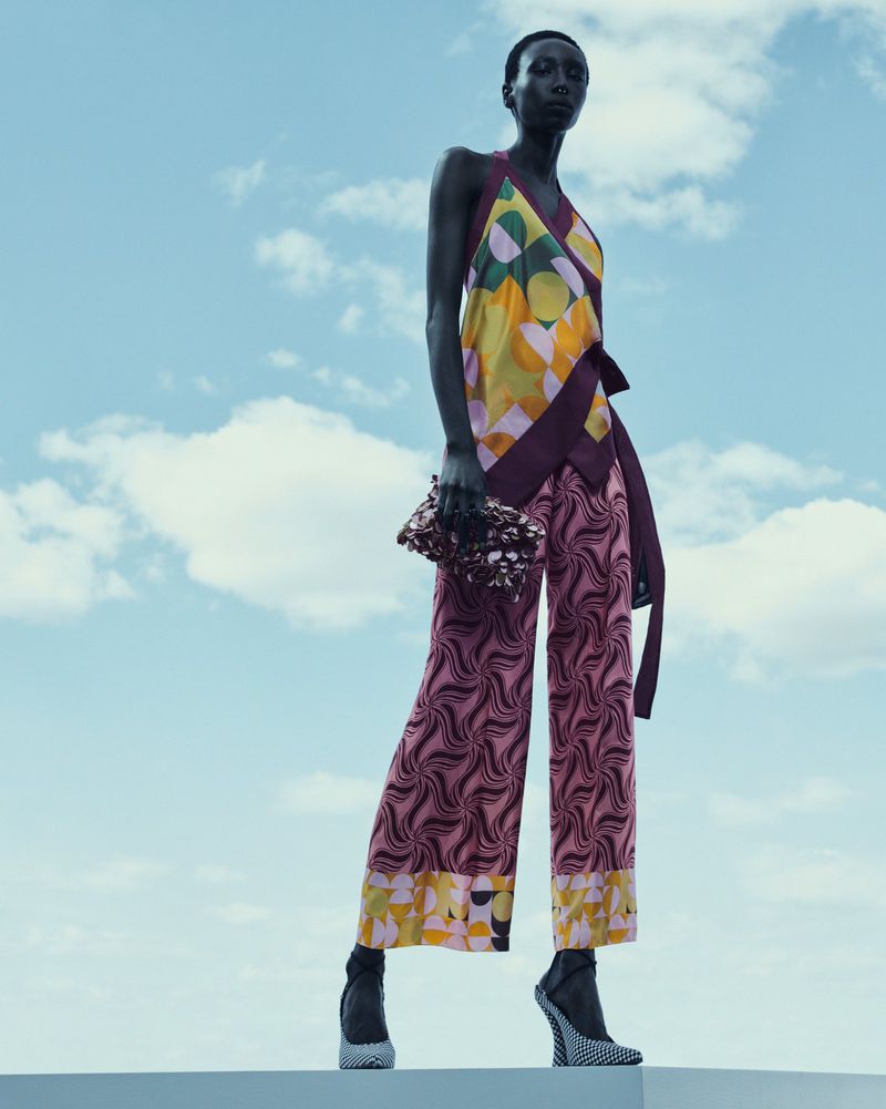 An adult female model poses in a geometric pattern top and pants with black heels and a clutch purse against a blue sky with clouds.
