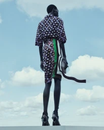 Model standing outdoors from a low angle against a partly cloudy blue sky, wearing a patterned top and skirt with a long tie detail.