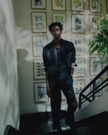 A young adult Black male is posing on stairs in a dark suit with botanical art prints on the wall in the background, shot with natural light.