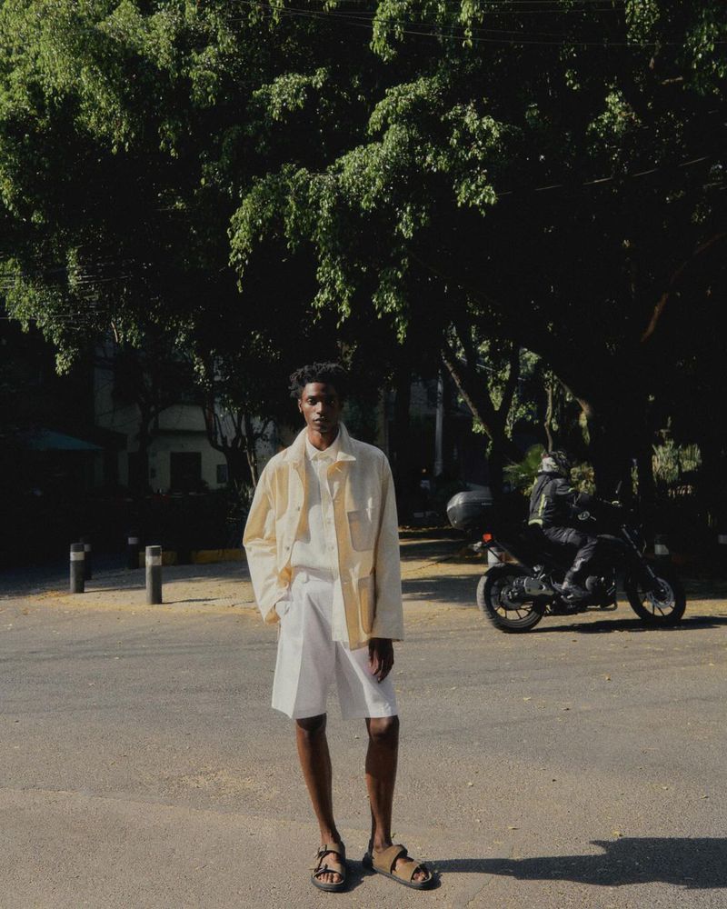 Full shot of a dark-skinned man in off-white jacket and shorts in an urban setting with a motorcycle visible in the background.