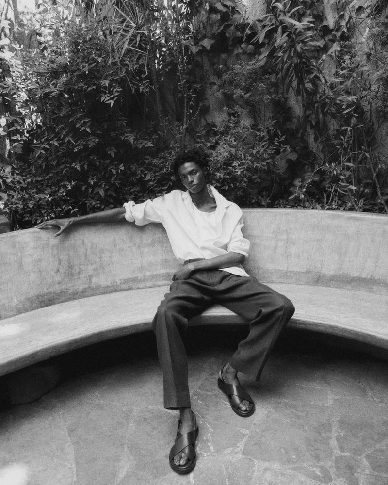 Portrait of a Black male model in an unbuttoned white shirt and trousers, sitting on a curved bench surrounded by green foliage.