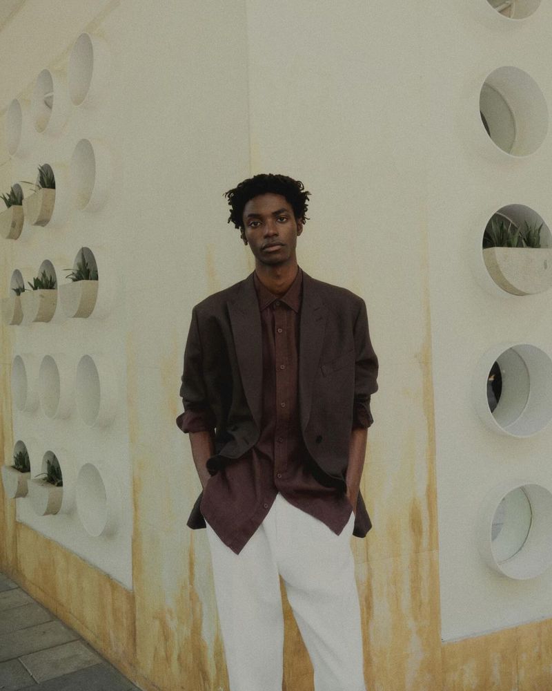 A young Black male poses in a brown blazer and white pants against a white wall with circular inset planters in an indoor setting.