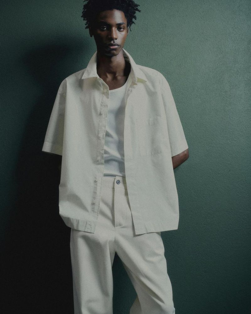 Adult male poses indoors wearing an open white short sleeve shirt, a white t-shirt, and white pants against a textured green wall.