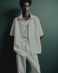 Young Black male model wearing a white button-down shirt and light trousers against a dark green textured wall.
