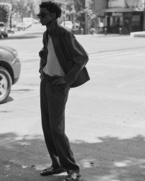 Black and white portrait of a young man wearing sunglasses and a jacket standing on a sunny urban street.