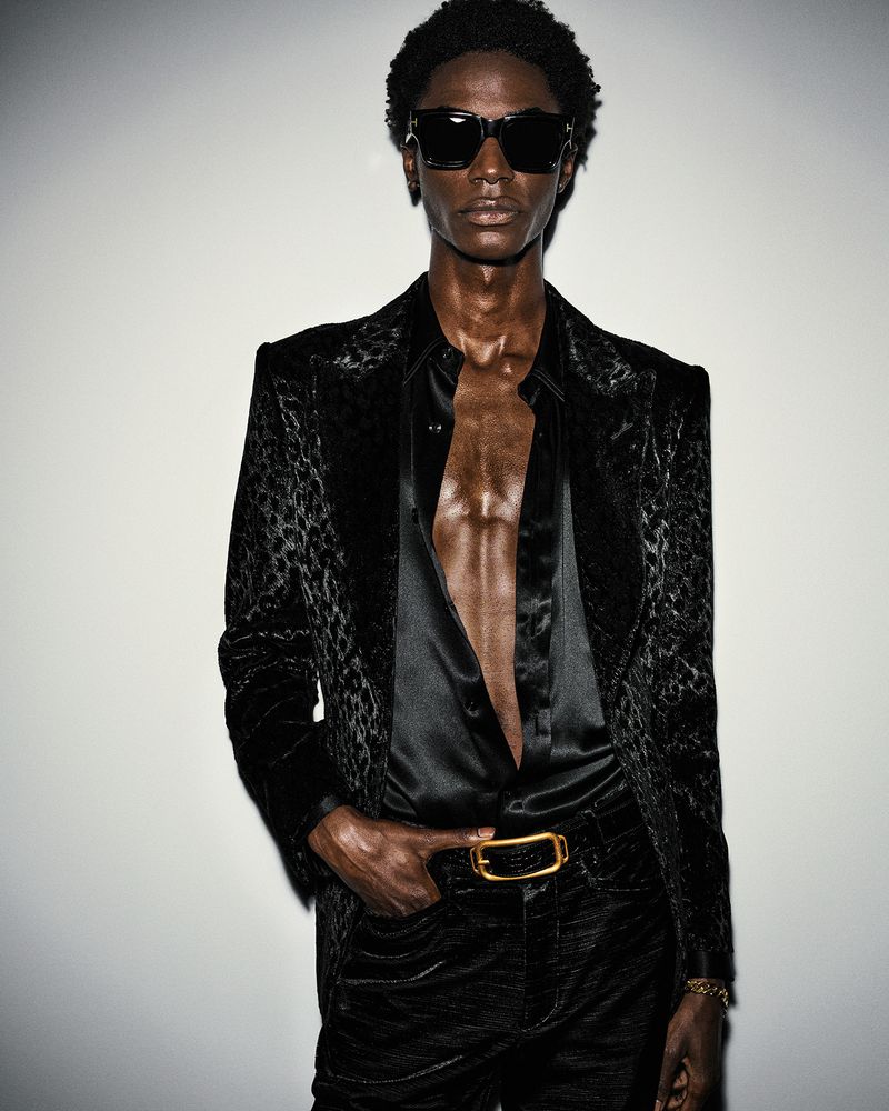 Dark-skinned male model poses in a patterned black velvet blazer and unbuttoned black shirt, accessorized with sunglasses and a gold belt.