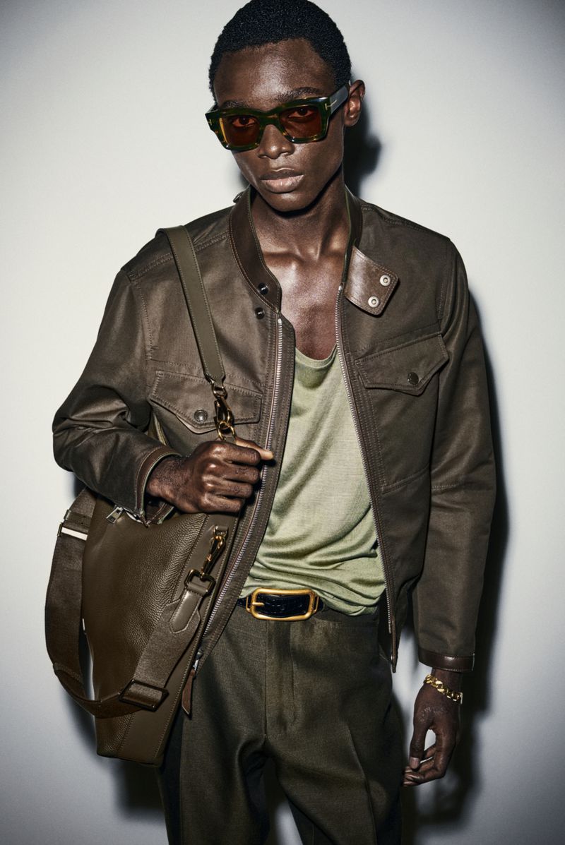 Dark-skinned male model poses in olive-green jacket with matching pants and bag, wearing sunglasses and gold jewelry in studio.