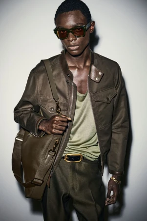 Dark-skinned male model poses in olive-green jacket with matching pants and bag, wearing sunglasses and gold jewelry in studio.